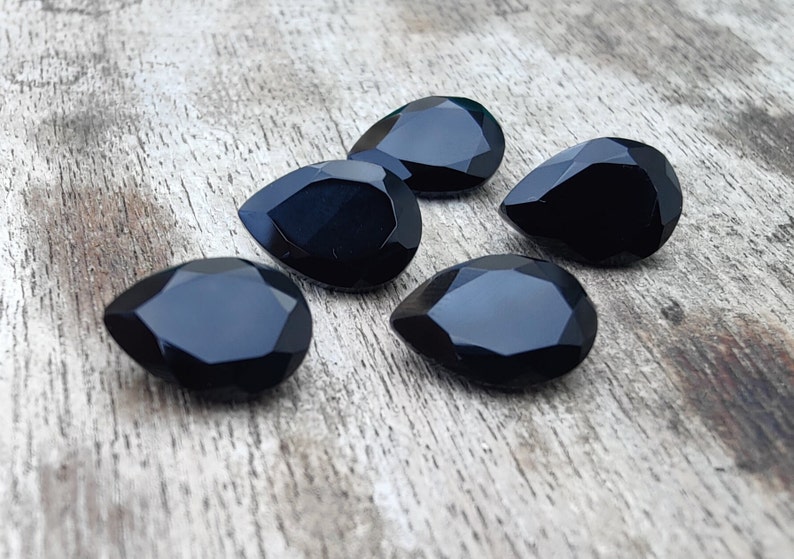 May include: Five black teardrop-shaped faceted gemstones on a wooden surface.