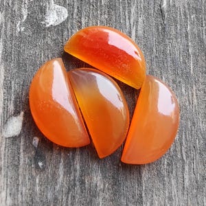 Puede incluir: Cuatro cabujones de piedra de cornalina naranja, cada uno cortado en forma de semicírculo. Los cabujones están pulidos y tienen un acabado liso y brillante.