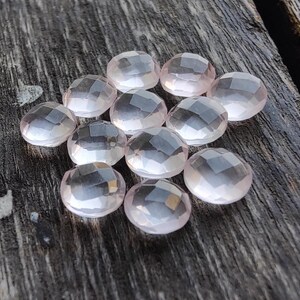 May include: A group of 12 faceted rose quartz stones, each with a light pink hue and a smooth, polished finish. The stones are arranged in a loose cluster on a wooden surface.