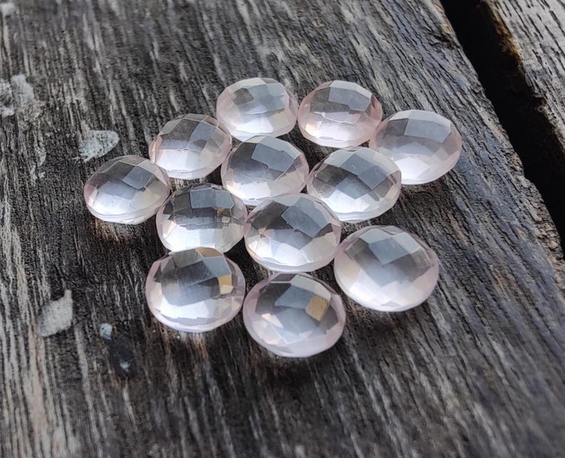 May include: A group of 12 faceted rose quartz stones, each with a light pink hue and a smooth, polished finish. The stones are arranged in a loose cluster on a wooden surface.