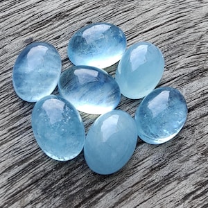 Può includere: Sette pietre preziose di acquamarina ovali e lisce, in diverse tonalità di azzurro chiaro. Le pietre sono disposte a grappolo su una superficie di legno.