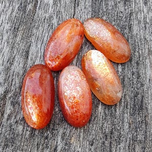K&ouml;nnte beinhalten: F&uuml;nf ovale Sonnensten-Cabochons mit schimmerndem, irisierendem Finish. Die Steine sind in Orange- und Rott&ouml;nen gehalten und haben einen dezenten Glanz.