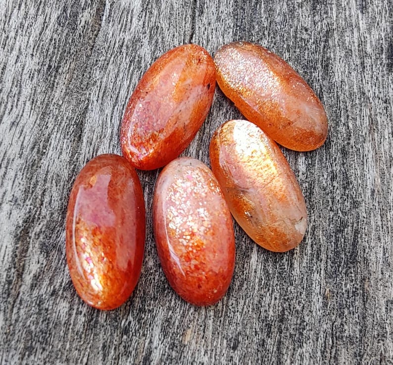 K&ouml;nnte beinhalten: F&uuml;nf ovale Sonnensten-Cabochons mit schimmerndem, irisierendem Finish. Die Steine sind in Orange- und Rott&ouml;nen gehalten und haben einen dezenten Glanz.