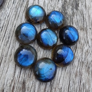 Puede incluir: Ocho piedras preciosas redondas de labradorita azul iridiscente con un brillo azul oscuro y verde. Las piedras están dispuestas en círculo sobre una superficie de madera.