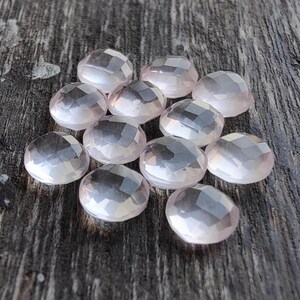 May include: A collection of 11 faceted rose quartz stones, each with a light pink hue and a smooth, polished finish. The stones are arranged on a wooden surface.