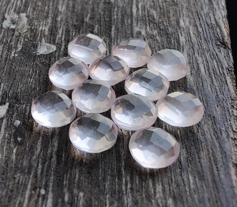 May include: A collection of 11 faceted rose quartz stones, each with a light pink hue and a smooth, polished finish. The stones are arranged on a wooden surface.
