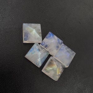 May include: Five square-cut moonstone gemstones with iridescent blue and white hues. The gemstones are arranged on a dark surface, showcasing their polished facets and natural color variations. These stones are ideal for jewelry making.