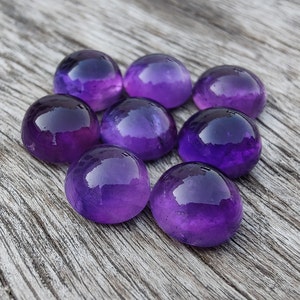 May include: Eight round, polished amethyst gemstones in various shades of purple. The gemstones are arranged in a circular pattern on a wooden surface.