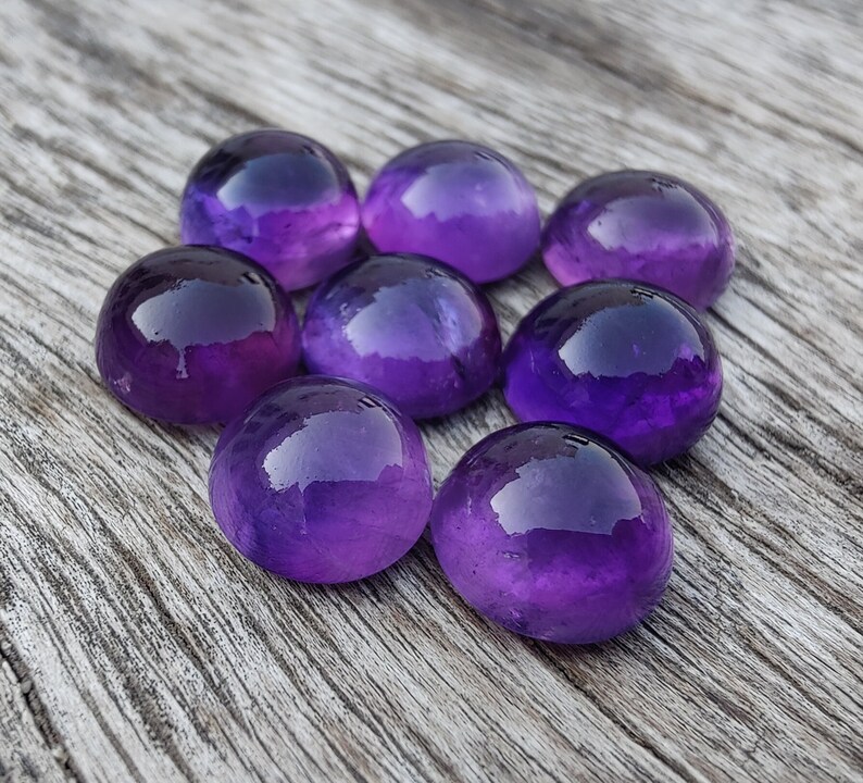 May include: Eight round, polished amethyst gemstones in various shades of purple. The gemstones are arranged in a circular pattern on a wooden surface.
