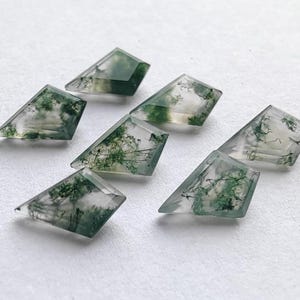 May include: Seven faceted moss agate gemstones. Each gemstone is a unique trapezoid shape with a clear base and green, tree-like inclusions. The gemstones are arranged on a white surface, showcasing their natural beauty and intricate patterns.