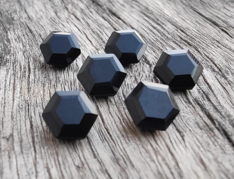 May include: Six black faceted glass beads, each with a unique geometric shape, arranged on a wooden surface.