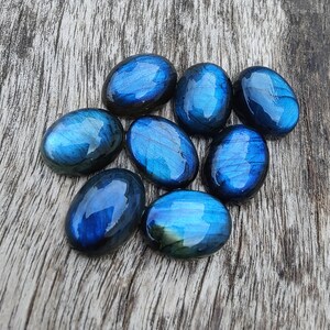 May include: A collection of eight oval-shaped labradorite stones, each with a unique iridescent blue and green sheen. The stones are arranged on a wooden surface.