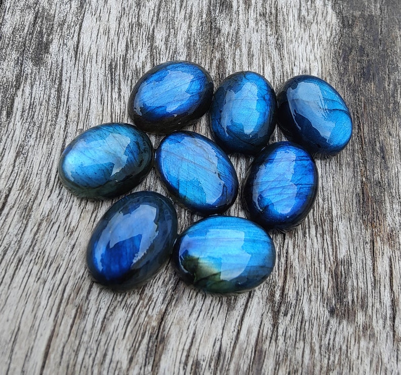 May include: A collection of eight oval-shaped labradorite stones, each with a unique iridescent blue and green sheen. The stones are arranged on a wooden surface.