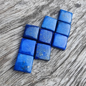 Puede incluir: Una colección de nueve pequeñas piedras cuadradas de lapislázuli azul con motas doradas. Las piedras están dispuestas en forma escalonada, creando una línea diagonal.