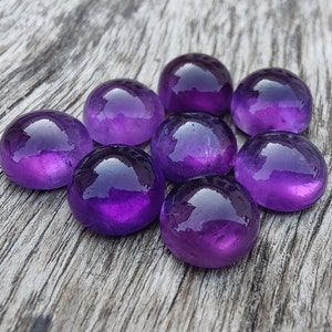 May include: Eight purple amethyst gemstones, each with a smooth, rounded surface, are arranged in a circular pattern on a weathered wooden surface.