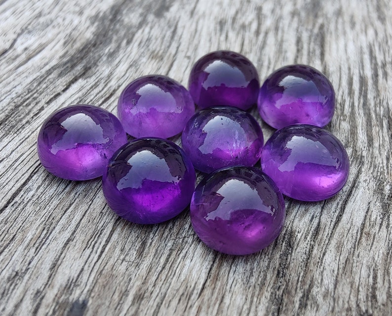 May include: Eight purple amethyst gemstones, each with a smooth, rounded surface, are arranged in a circular pattern on a weathered wooden surface.