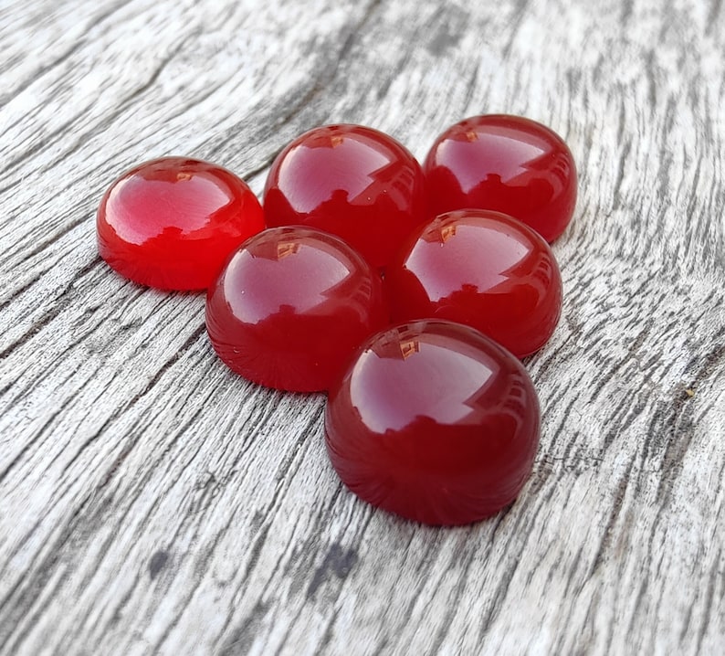 K&ouml;nnte beinhalten: Sechs rote Cabochon-Edelsteine auf einer Holzoberfl&auml;che. Die Edelsteine sind rund und haben eine glatte, polierte Oberfl&auml;che.