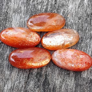 K&ouml;nnte beinhalten: Sechs ovale, getrommelte Sunstone-Steine mit schimmerndem, irisierendem Finish. Die Steine sind in einem Mix aus Orange- und Rott&ouml;nen mit einem metallischen Schimmer.
