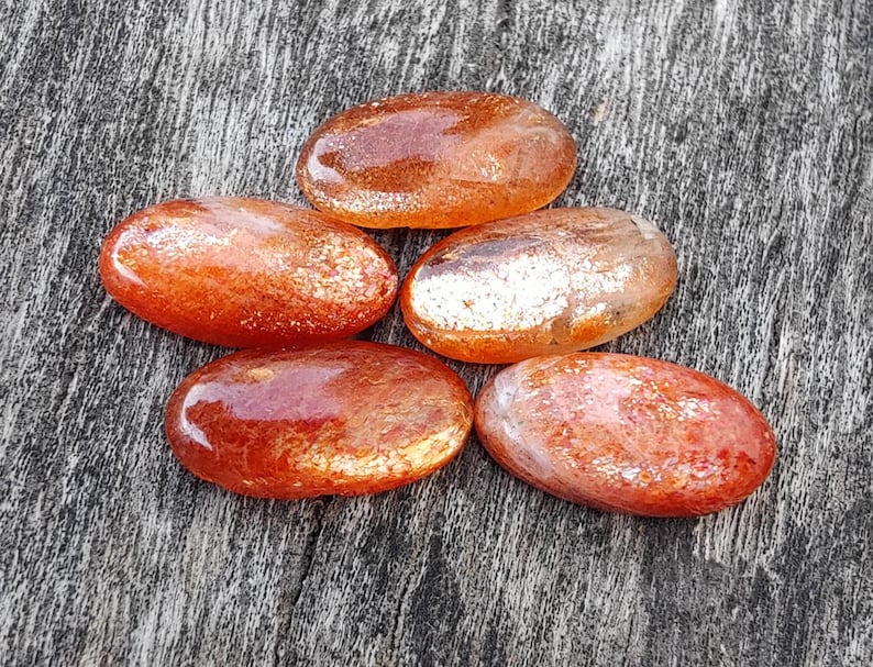 K&ouml;nnte beinhalten: Sechs ovale, getrommelte Sunstone-Steine mit schimmerndem, irisierendem Finish. Die Steine sind in einem Mix aus Orange- und Rott&ouml;nen mit einem metallischen Schimmer.