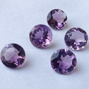 Puede incluir: Cinco piedras preciosas de amatista redondas y facetadas en varios tonos de púrpura. Las piedras preciosas están dispuestas en semicírculo sobre un fondo blanco.