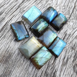 Könnte beinhalten: Eine Sammlung von acht schillernden Labradorit-Cabochons in verschiedenen Blautönen, Grüntönen und Goldtönen. Die Steine sind in rechteckiger Form geschliffen und haben eine glatte, polierte Oberfläche.