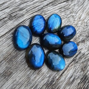 May include: Eight smooth, oval-shaped labradorite stones in shades of blue and green. The stones are arranged in a circular pattern on a wooden surface.