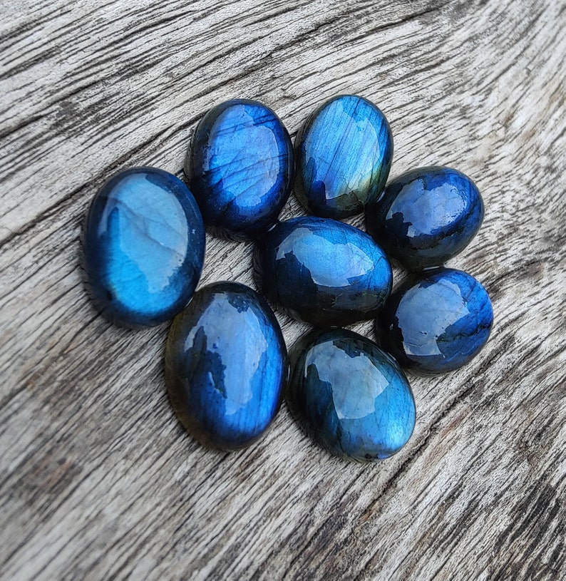 May include: Eight smooth, oval-shaped labradorite stones in shades of blue and green. The stones are arranged in a circular pattern on a wooden surface.