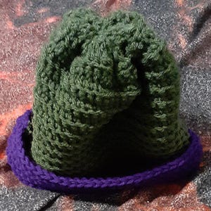 May include: A handmade crochet beanie featuring a textured olive green crown and a contrasting solid purple brim. The hat is constructed with a dense, repeating stitch pattern that creates a thick, warm appearance with a folded edge design.