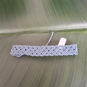 May include: A dusty blue macrame bracelet featuring a repeating diamond-shaped knot pattern. The bracelet has a flat, woven construction with a small, blank white rectangular tag attached to one side and a single loose cord extending from the top edge.