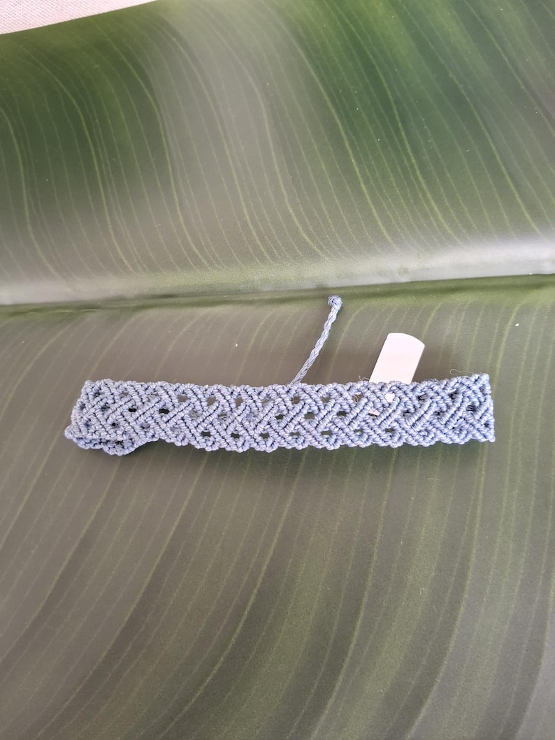 May include: A dusty blue macrame bracelet featuring a repeating diamond-shaped knot pattern. The bracelet has a flat, woven construction with a small, blank white rectangular tag attached to one side and a single loose cord extending from the top edge.