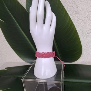 May include: A pink macrame bracelet featuring an intricate woven pattern of interconnected loops. The bracelet is secured with a thin cord tie and is displayed on a white hand-shaped stand. The design consists of repeating knot work.