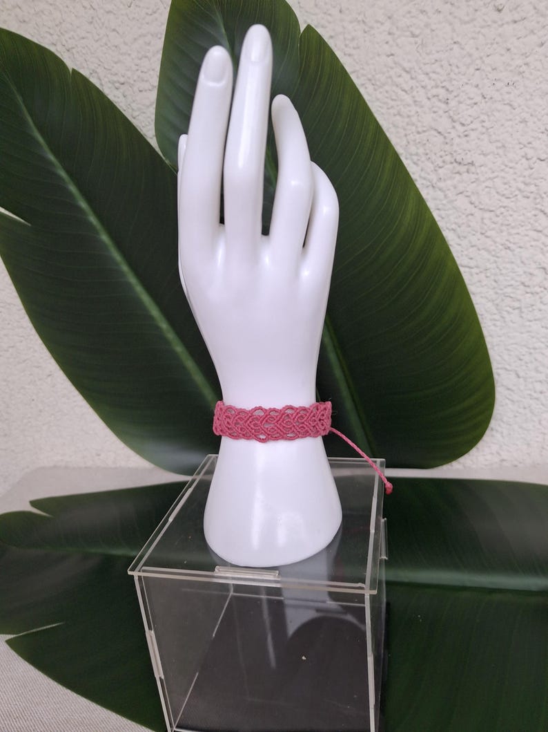 May include: A pink macrame bracelet featuring an intricate woven pattern of interconnected loops. The bracelet is secured with a thin cord tie and is displayed on a white hand-shaped stand. The design consists of repeating knot work.