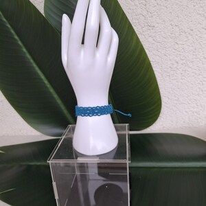 May include: A teal blue woven macrame bracelet featuring a repeating intricate knot pattern. The bracelet is secured with thin matching thread ties and is displayed on a white mannequin hand stand resting on a clear acrylic box.