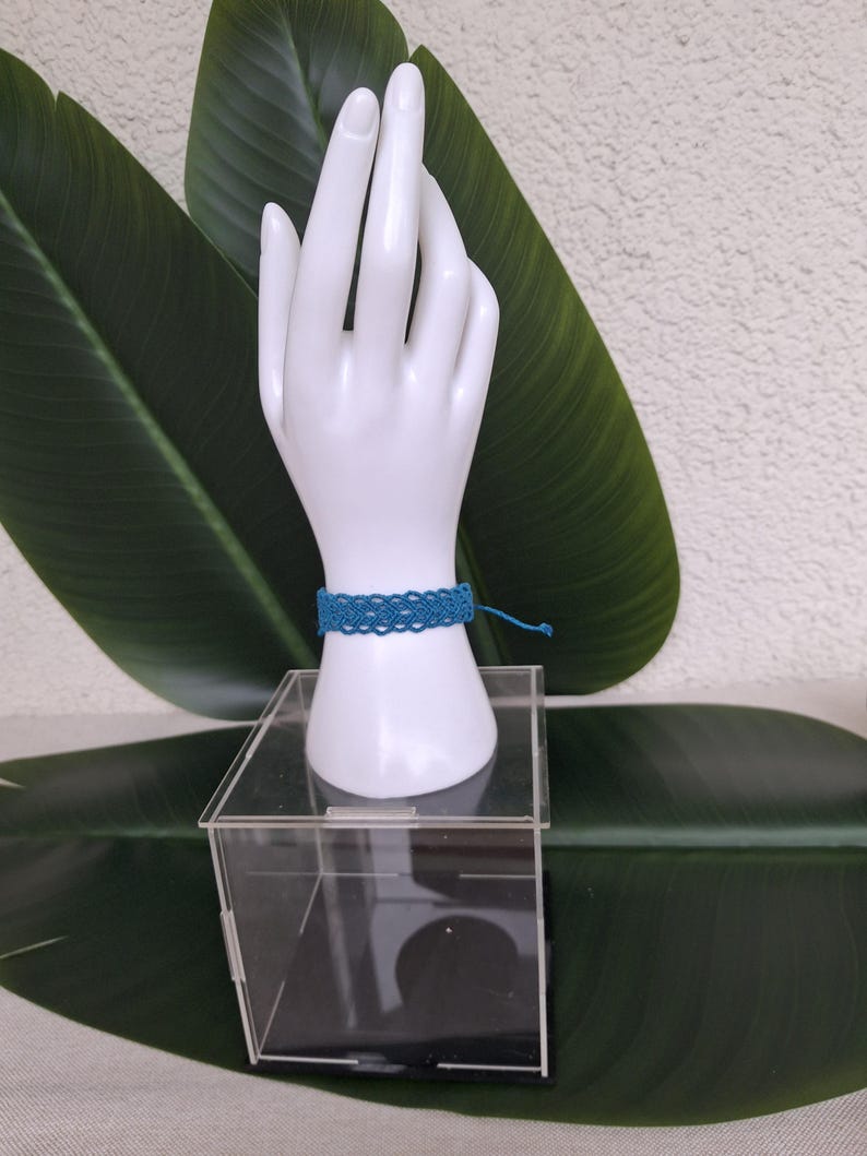 May include: A teal blue woven macrame bracelet featuring a repeating intricate knot pattern. The bracelet is secured with thin matching thread ties and is displayed on a white mannequin hand stand resting on a clear acrylic box.