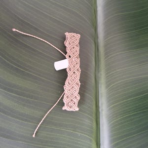 May include: A light brown macrame bracelet featuring an intricate woven pattern of knotted cord. The bracelet includes two long, thin adjustable cord ends and a small, rectangular white tag attached to the side of the woven section.