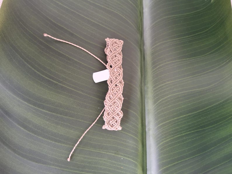 May include: A light brown macrame bracelet featuring an intricate woven pattern of knotted cord. The bracelet includes two long, thin adjustable cord ends and a small, rectangular white tag attached to the side of the woven section.