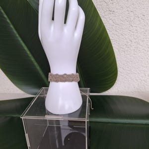 May include: A beige macrame bracelet featuring an intricate woven pattern of knotted cord. The bracelet is displayed on a white mannequin hand stand, which is positioned on top of a clear acrylic box. The design consists of repeating loops.