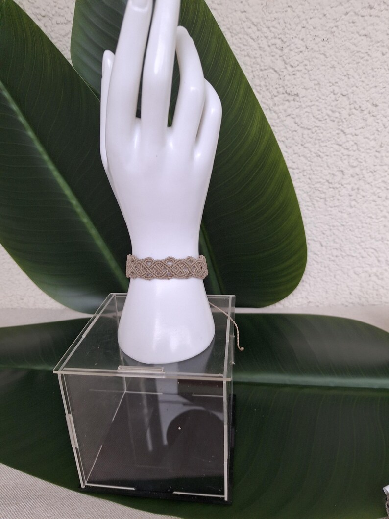 May include: A beige macrame bracelet featuring an intricate woven pattern of knotted cord. The bracelet is displayed on a white mannequin hand stand, which is positioned on top of a clear acrylic box. The design consists of repeating loops.