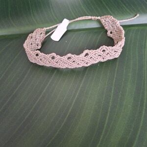 May include: A light brown macrame bracelet featuring an intricate woven pattern with scalloped edges and a small white rectangular tag attached to the cord. The bracelet is constructed from thin, knotted thread and includes adjustable tie closures.