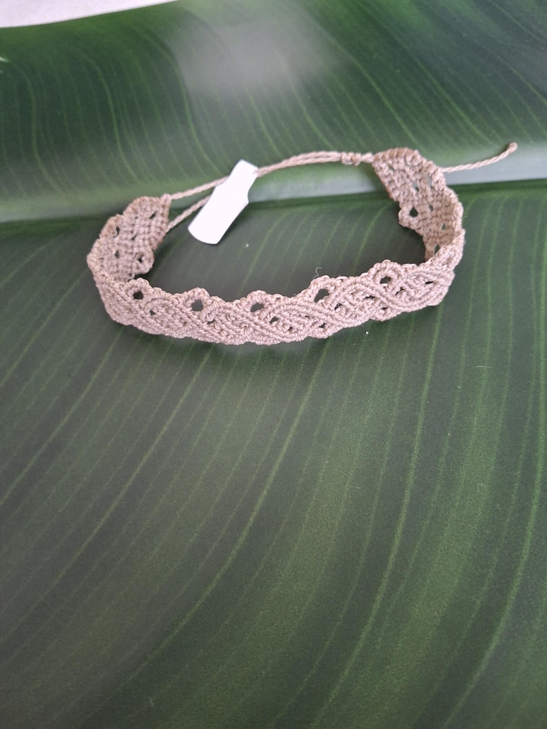 May include: A light brown macrame bracelet featuring an intricate woven pattern with scalloped edges and a small white rectangular tag attached to the cord. The bracelet is constructed from thin, knotted thread and includes adjustable tie closures.