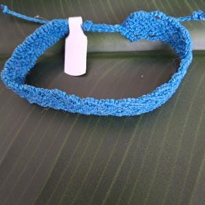 May include: A bright blue crocheted bracelet featuring a textured, woven pattern and thin tie-ends for closure. A small, blank white rectangular tag is attached to the band, which is constructed from a single continuous piece of blue fiber.