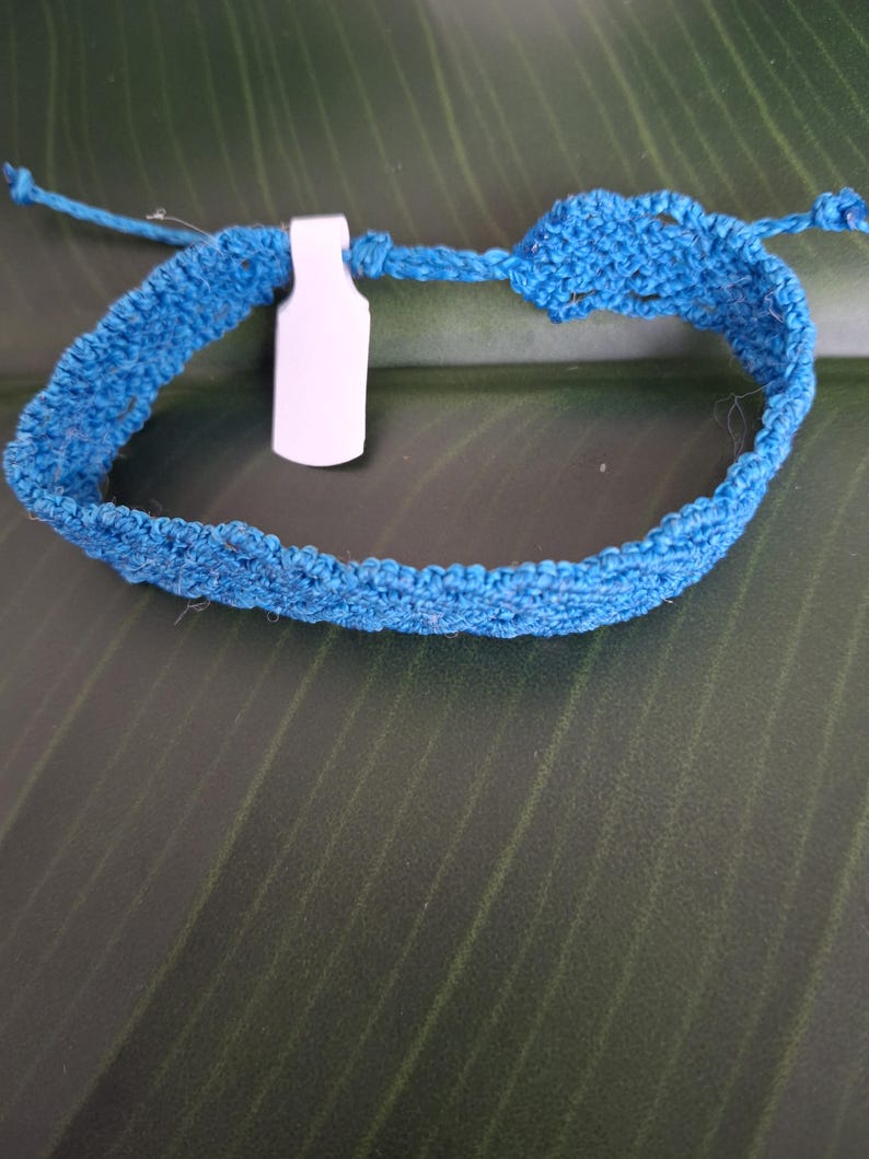 May include: A bright blue crocheted bracelet featuring a textured, woven pattern and thin tie-ends for closure. A small, blank white rectangular tag is attached to the band, which is constructed from a single continuous piece of blue fiber.