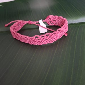 May include: A pink macrame bracelet featuring an intricate woven pattern with a scalloped edge. The bracelet includes thin adjustable cord ties and a small white rectangular tag attached to the center of the woven band.