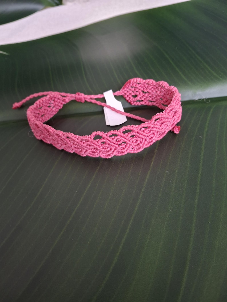 May include: A pink macrame bracelet featuring an intricate woven pattern with a scalloped edge. The bracelet includes thin adjustable cord ties and a small white rectangular tag attached to the center of the woven band.