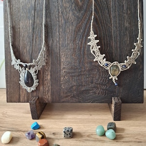 May include: Two macrame necklaces featuring intricate beige cord knotting and central gemstone pendants. The left necklace has a dark teardrop stone, while the right necklace features a round, iridescent stone with small blue bead accents.