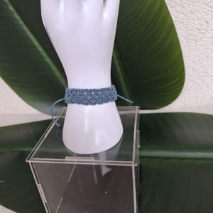 May include: A dusty blue macrame bracelet featuring an intricate woven pattern with thin cord ties on each side. The bracelet is displayed on a white mannequin hand form, showcasing the detailed knot work and the soft, muted blue color of the cord.