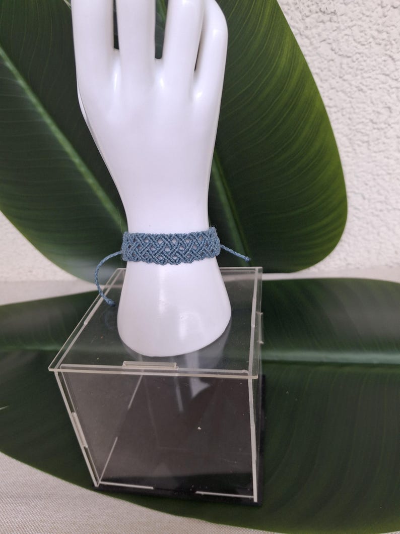May include: A dusty blue macrame bracelet featuring an intricate woven pattern with thin cord ties on each side. The bracelet is displayed on a white mannequin hand form, showcasing the detailed knot work and the soft, muted blue color of the cord.