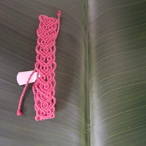 May include: A vibrant pink macrame bracelet featuring a repeating heart-shaped knot pattern. The bracelet is constructed from thin cord and includes two long, braided ties at the top for fastening. A small white rectangular tag is attached to the side.