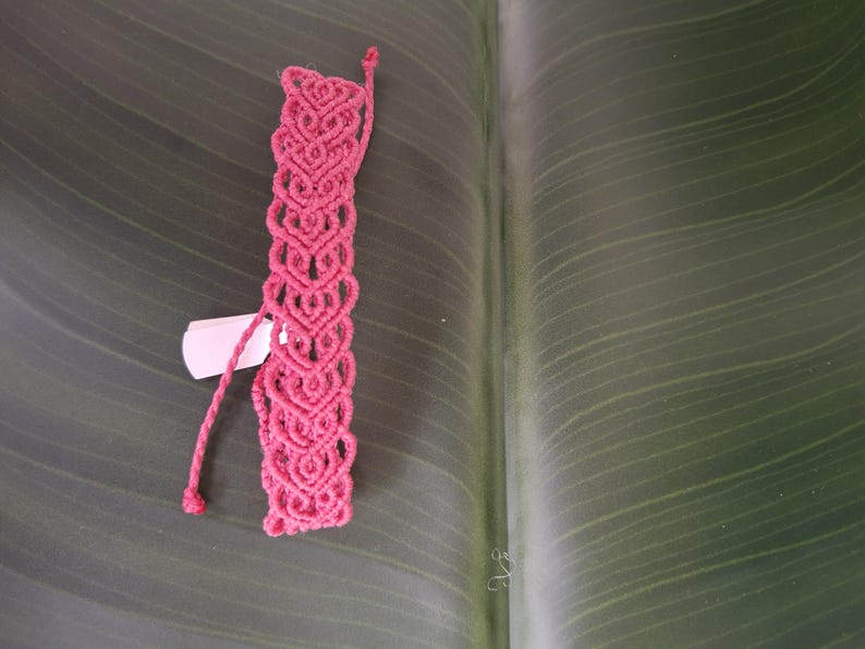 May include: A vibrant pink macrame bracelet featuring a repeating heart-shaped knot pattern. The bracelet is constructed from thin cord and includes two long, braided ties at the top for fastening. A small white rectangular tag is attached to the side.