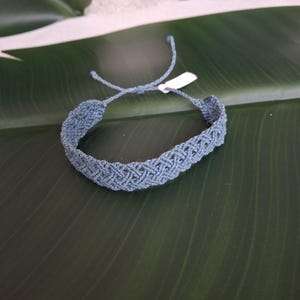 May include: A dusty blue macrame bracelet featuring an intricate woven chevron pattern. The bracelet is constructed from thin cord and includes adjustable tie closures with loose ends and a small white rectangular tag attached to one side.